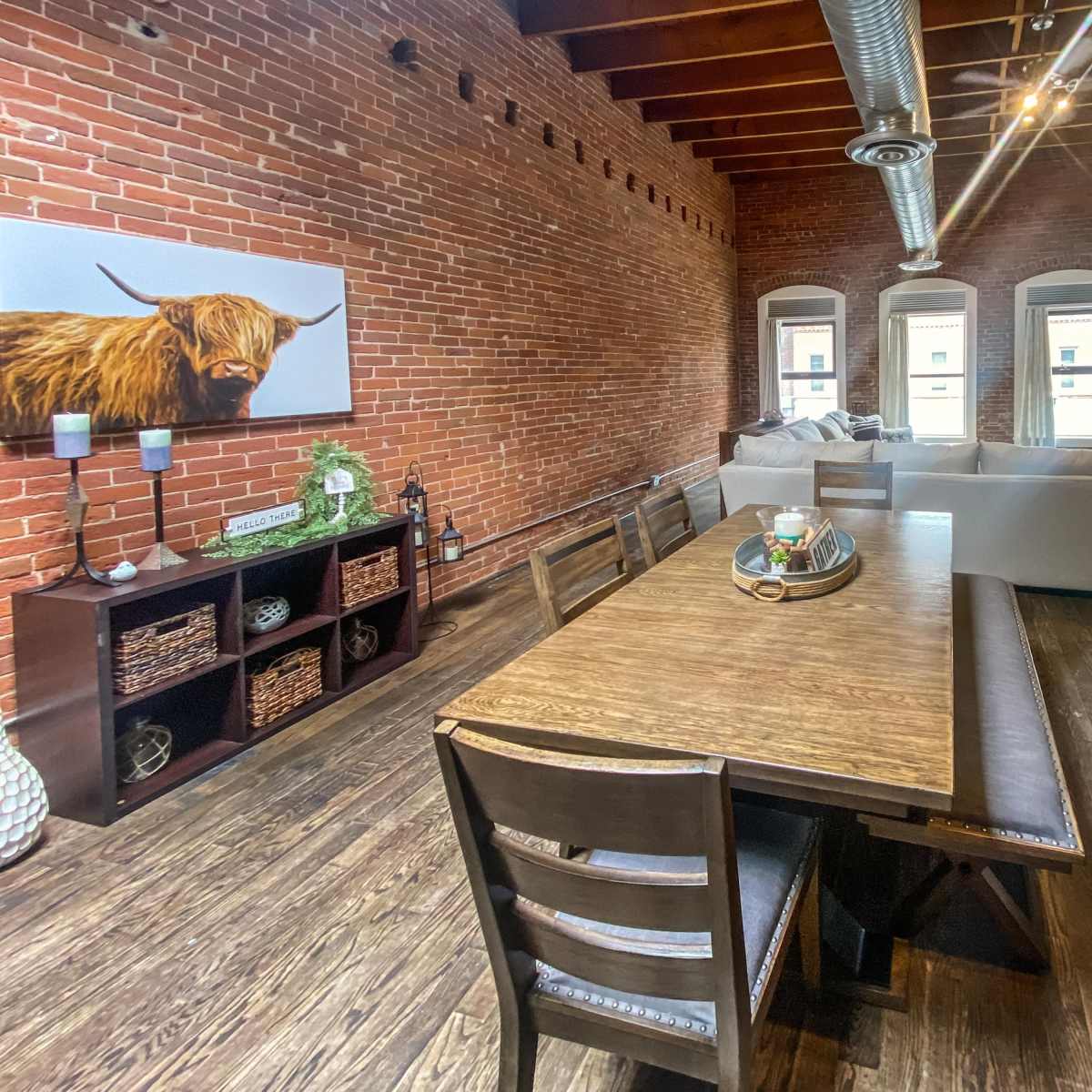 A spacious living and dining area featuring exposed brick walls, a wooden dining table with chairs, and a large sectional sofa near windows.