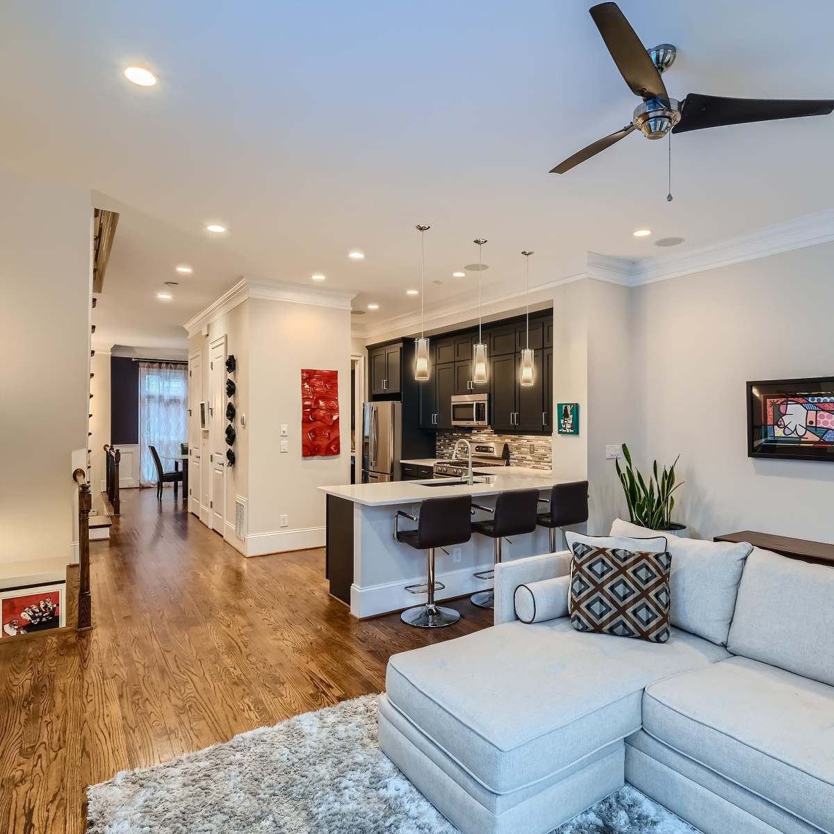 A modern living room with a sectional couch, a kitchen area featuring a breakfast bar, and hardwood floors leading to a hallway.