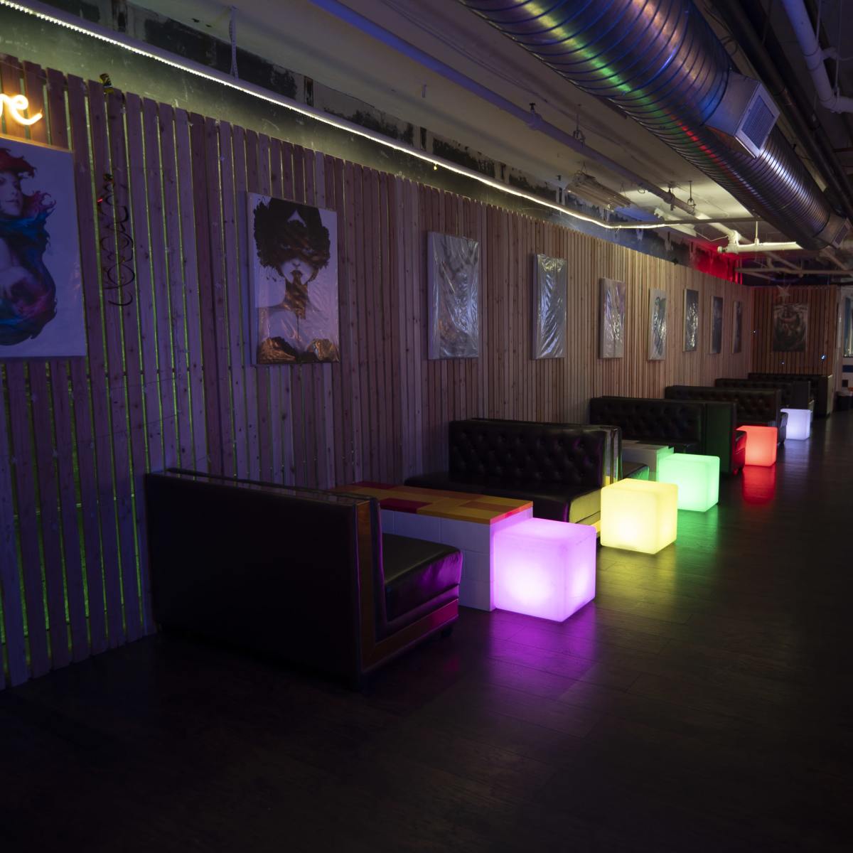 A dimly lit lounge area with seating and colorful illuminated tables along the walls decorated with art.