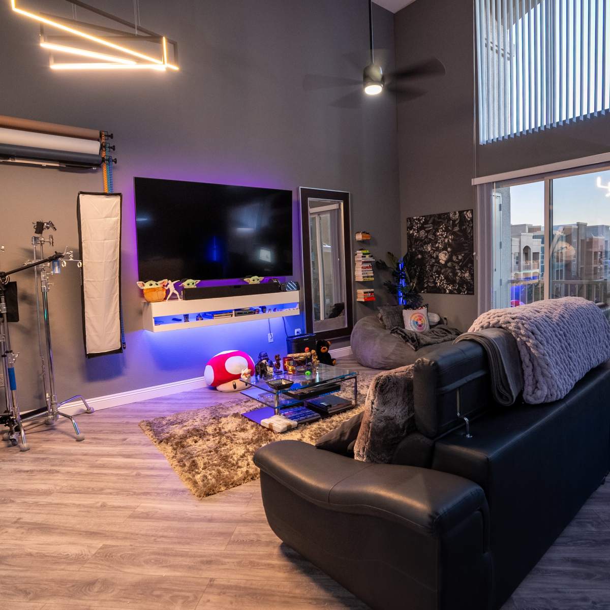 A modern living space with a dark gray wall, a sectional sofa, a large screen mounted on the wall, ambient lighting, and various equipment for video production.
