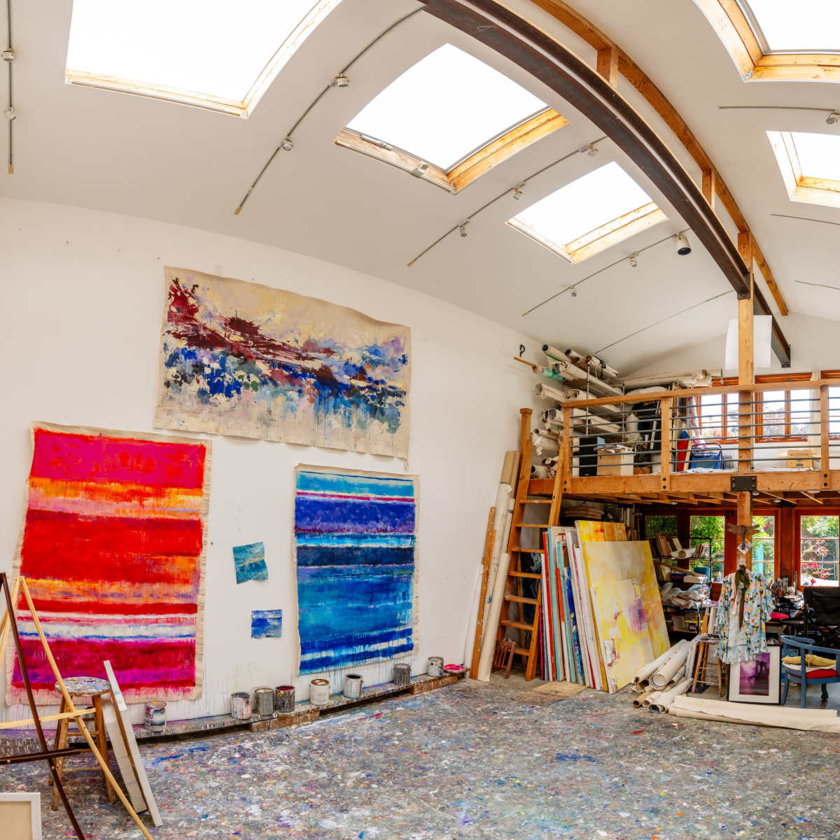 A spacious artist's studio with large skylights, colorful paintings on the walls, and scattered art supplies on the floor.