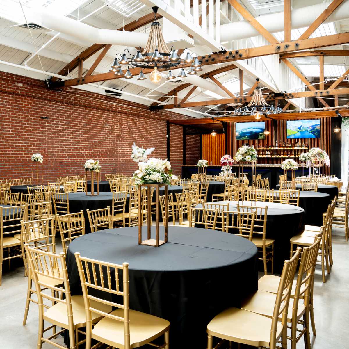 A spacious event venue with round tables covered in black linens and gold chairs, set against a backdrop of exposed red brick walls and wooden beams.