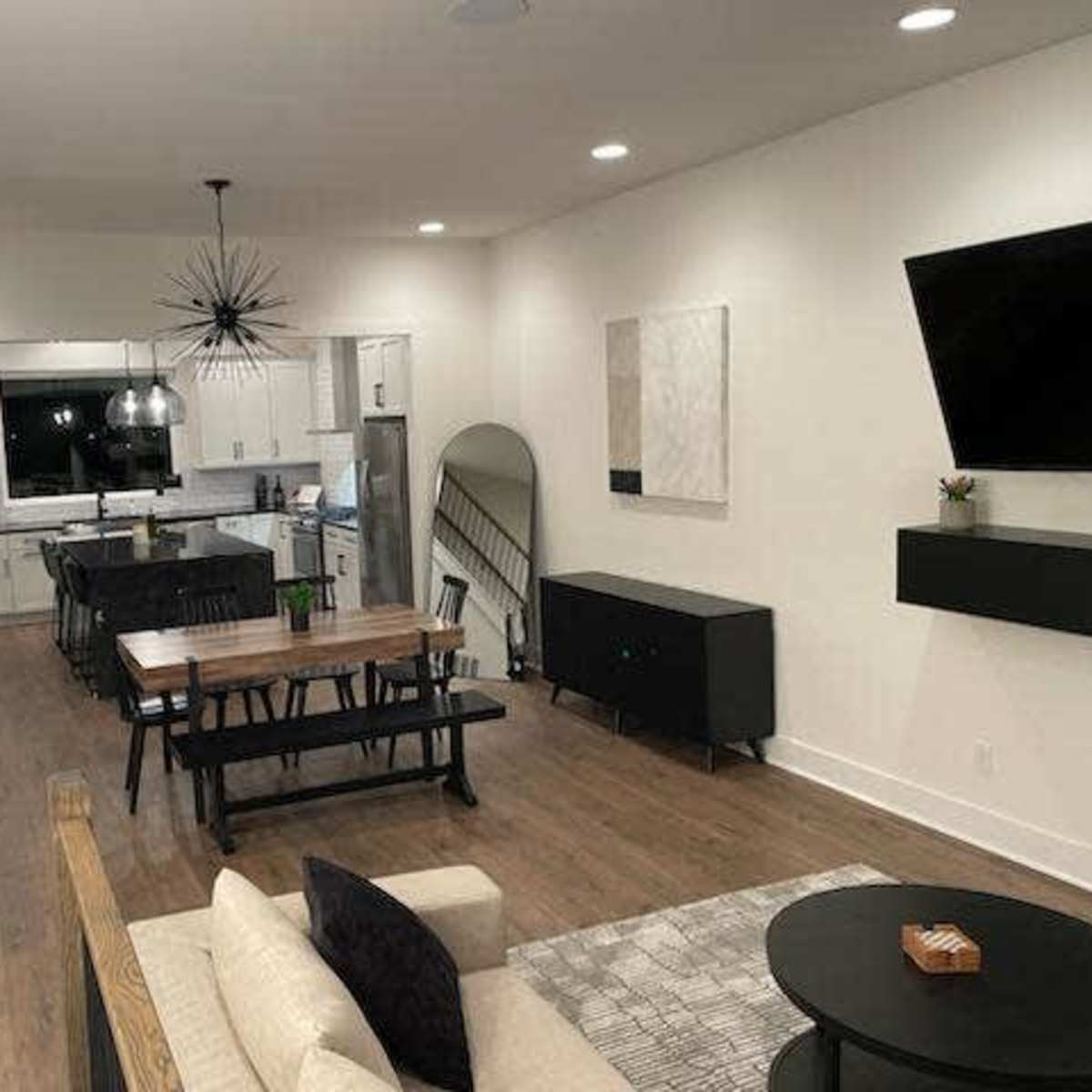 A modern living room connected to a kitchen and dining area, featuring a wooden floor, a large table, and contemporary lighting fixtures.