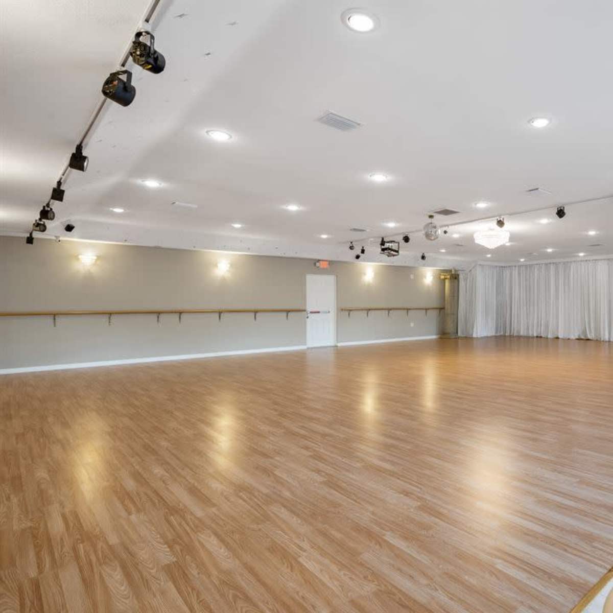 A spacious, well-lit dance studio with polished wooden floors and a mirrored wall, equipped with overhead lighting and a small refreshment area.