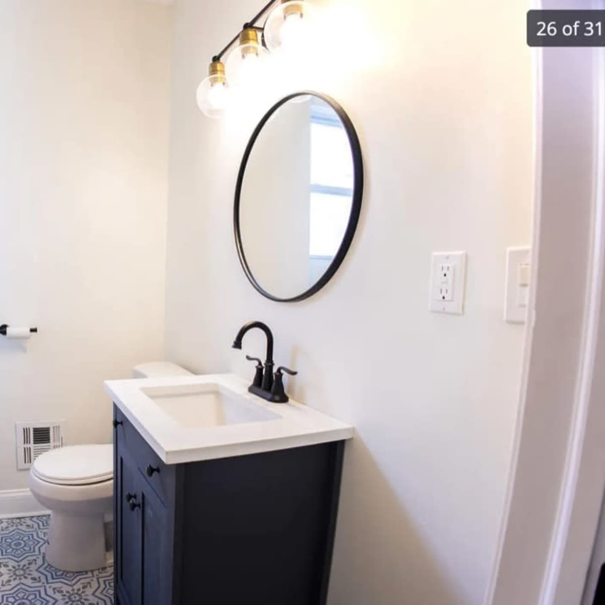 A small bathroom featuring a shower, toilet, and a sink with a dark cabinet, illuminated by three light bulbs and a round mirror above the sink.