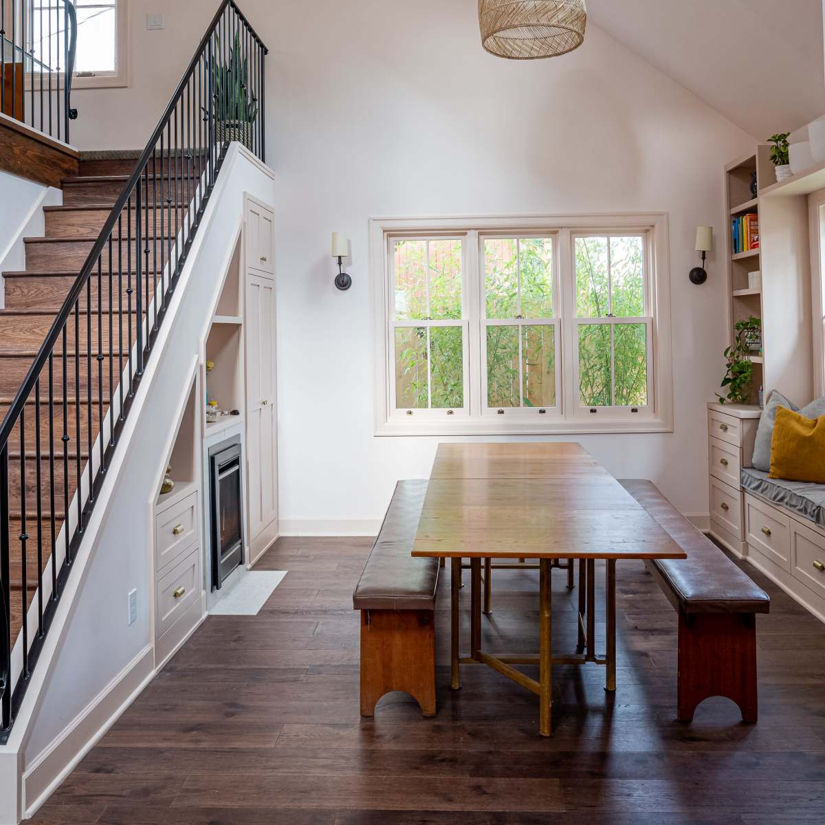 A dining area with a large wooden table and benches is located beneath a sloped ceiling, with a staircase nearby and large windows allowing natural light to enter.