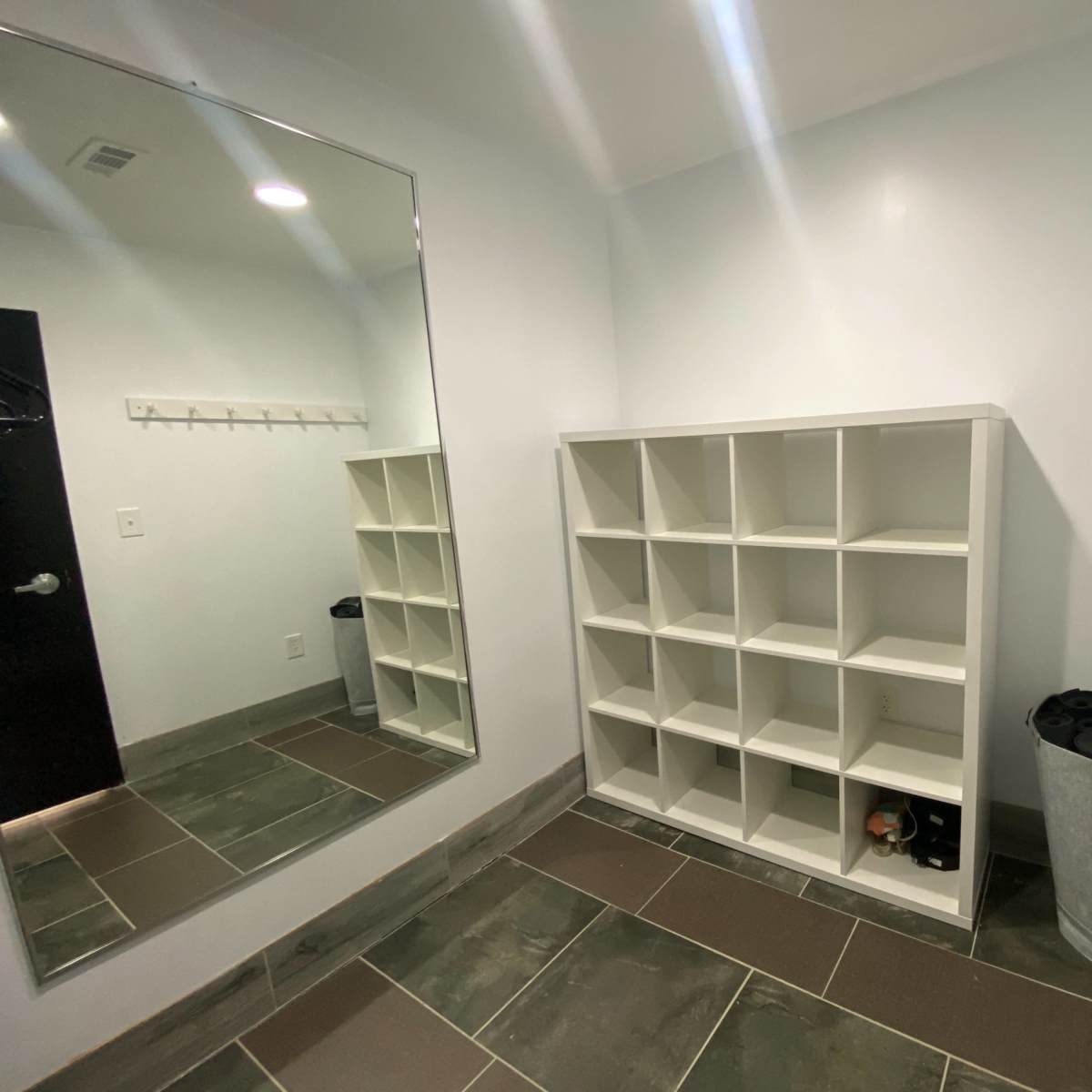 A minimalist entryway with a large mirror, a cubical shelving unit, a trash can, and a wall-mounted rack for hanging items.