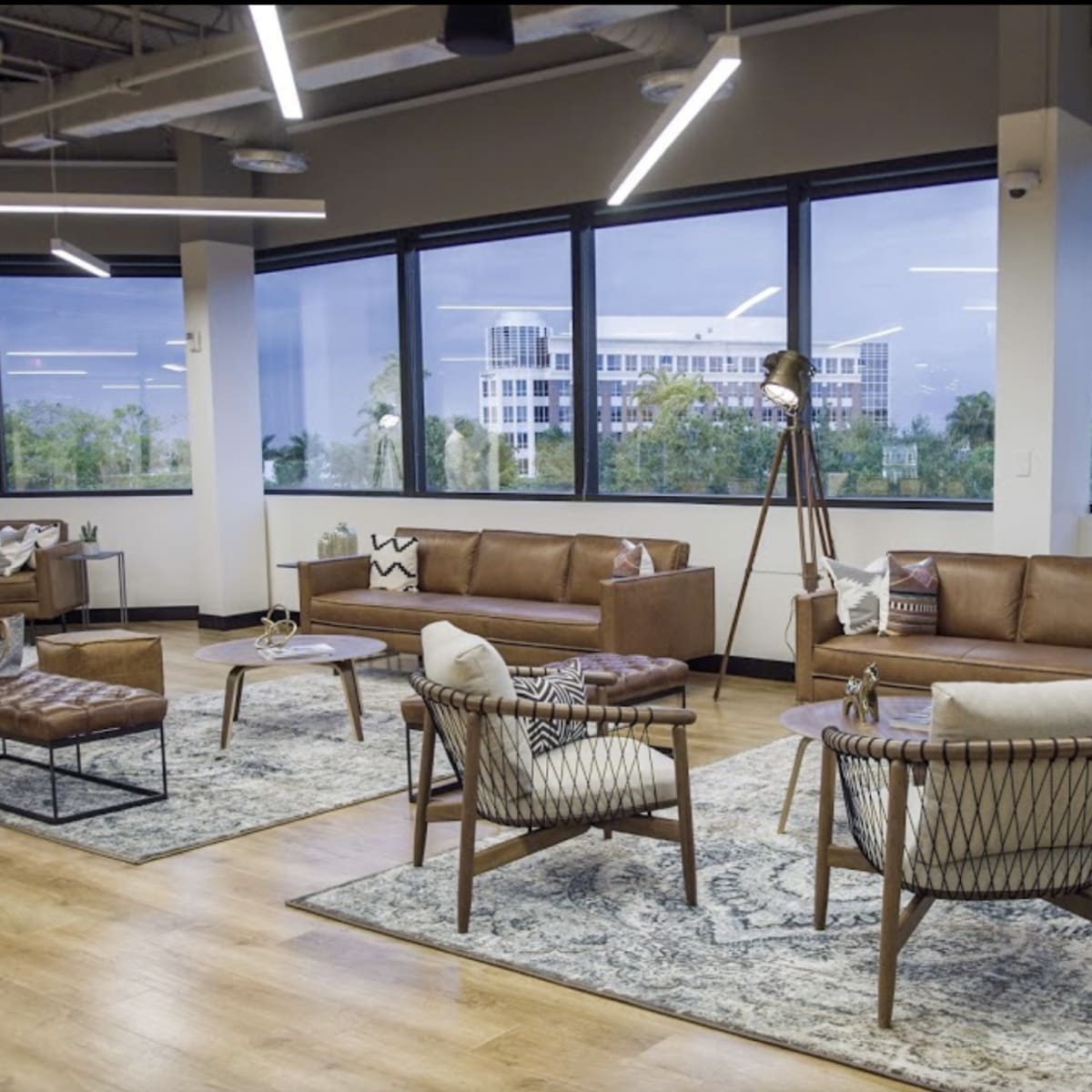 A modern office lounge area with brown leather sofas, woven chairs, a round coffee table, and large windows offering a view of the outdoors.