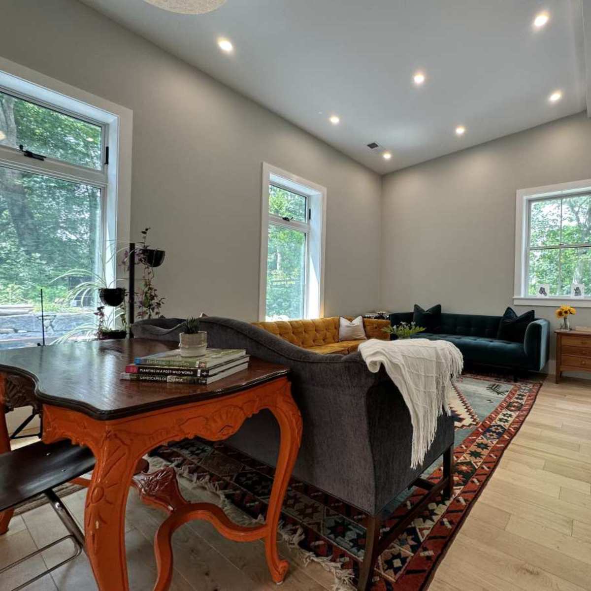 A bright living room featuring a gray sofa, a green couch, a wooden dining table, and large windows with views of greenery outside.