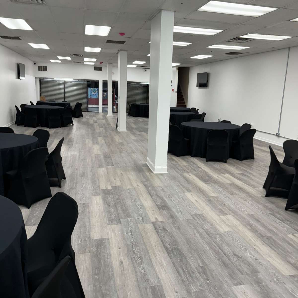 A spacious event room with black tablecloths covering round tables arranged throughout, and a light-colored wooden floor.