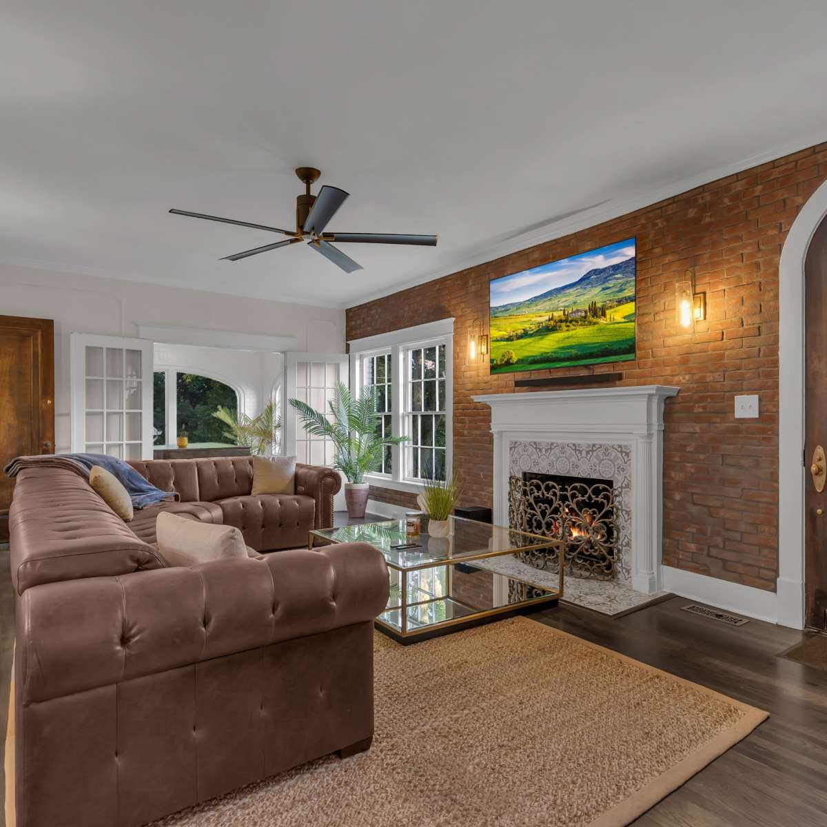 The living room features a brown sectional sofa, a glass coffee table, a TV mounted above a white fireplace, and a large wooden front door.