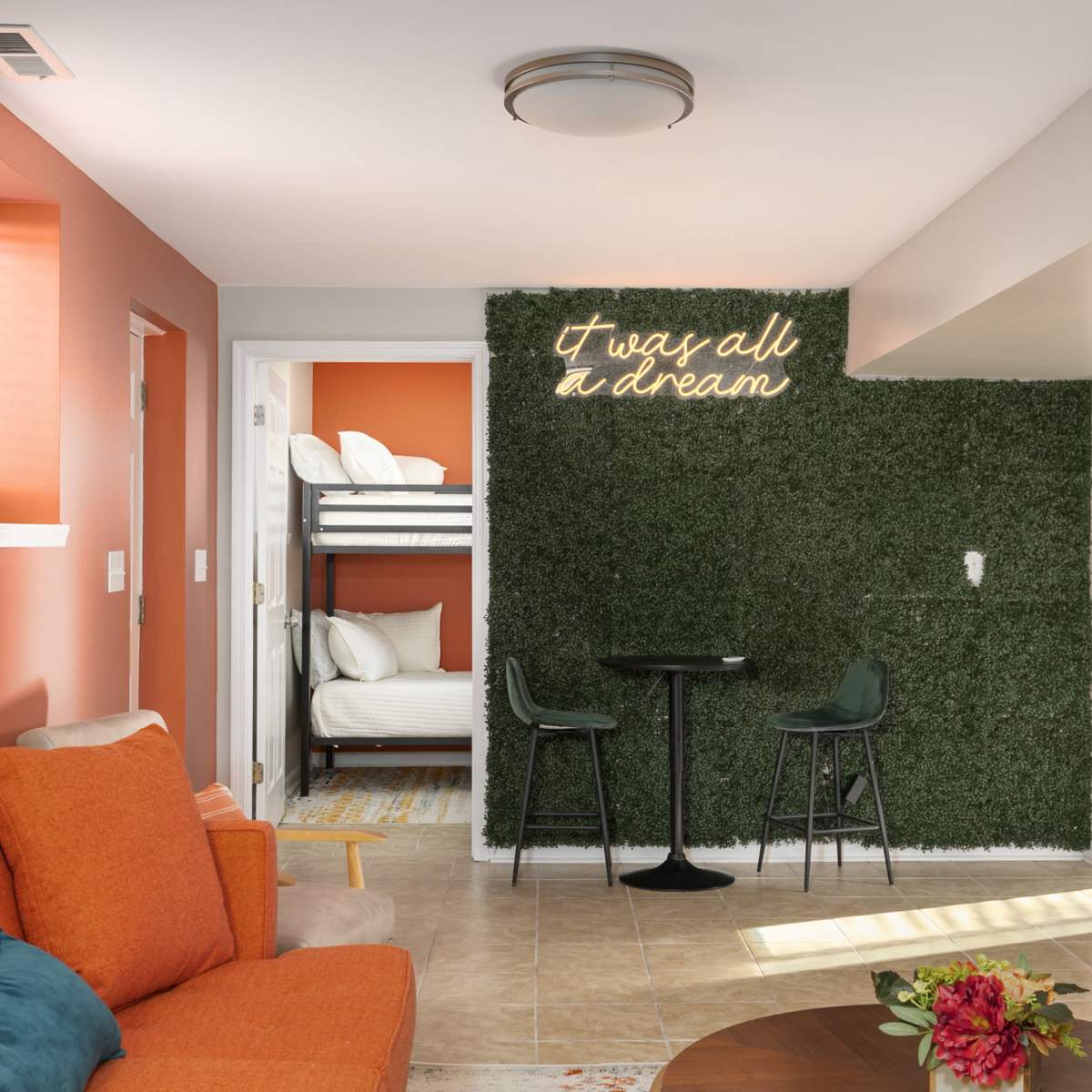 A well-lit modern living space features an orange sofa, a small dining table, and a green wall adorned with the phrase "I was all a dream."