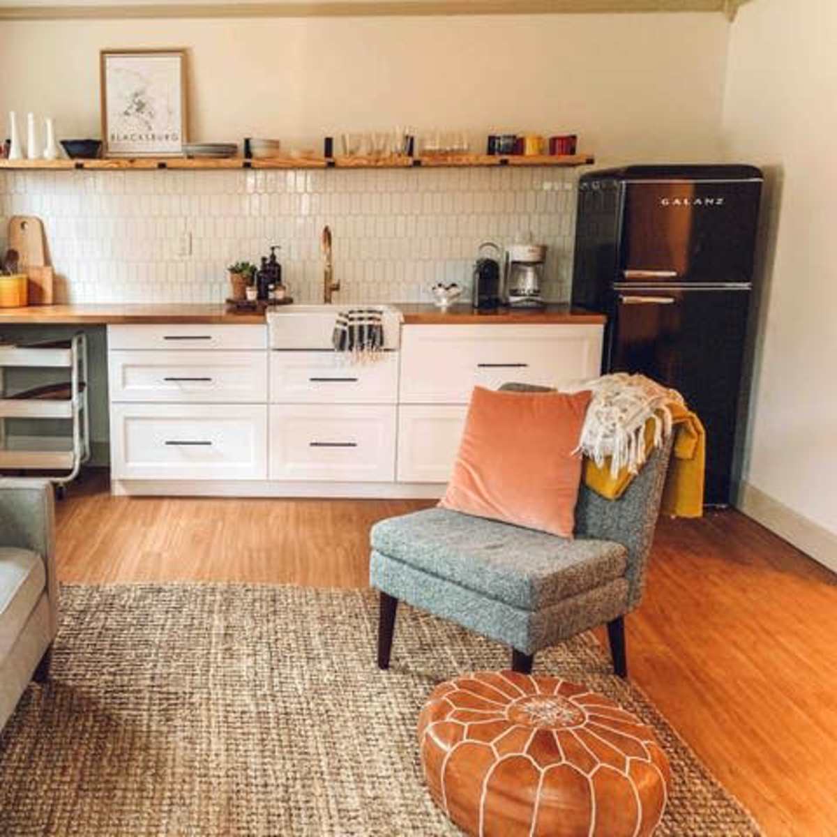 A modern kitchen and living area feature a white cabinetry, wooden countertop, and a gray couch with orange pillows, set on a woven rug.