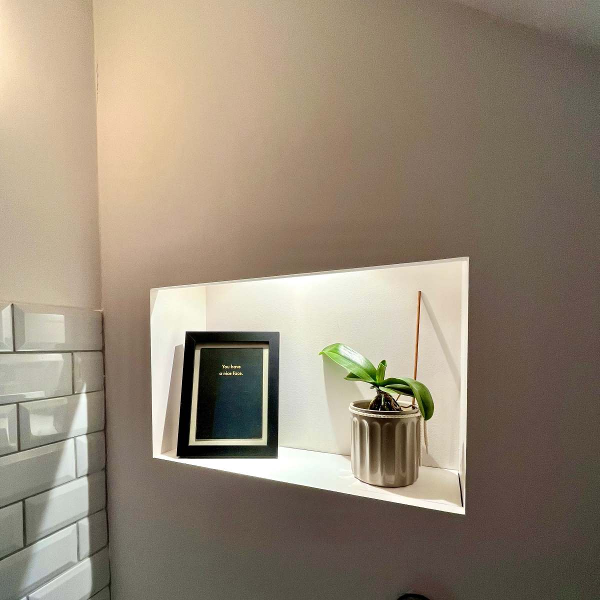 A lit alcove with a framed picture and a potted plant on a shelf against a white wall.