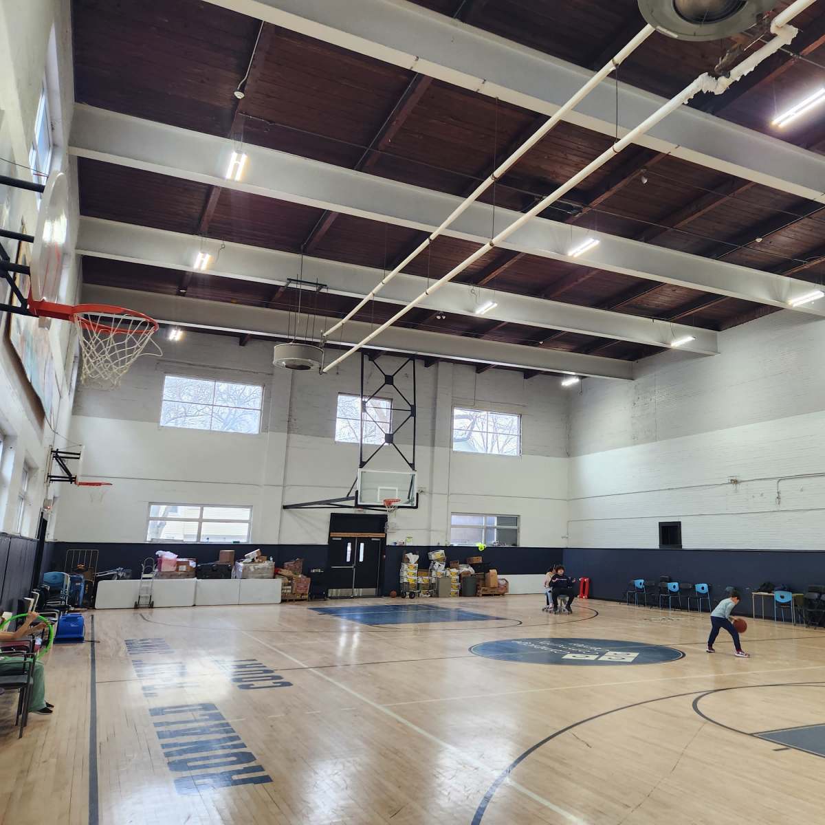A basketball court is shown with a high ceiling, featuring a hoop and two children playing while boxes and chairs are arranged along the walls.
