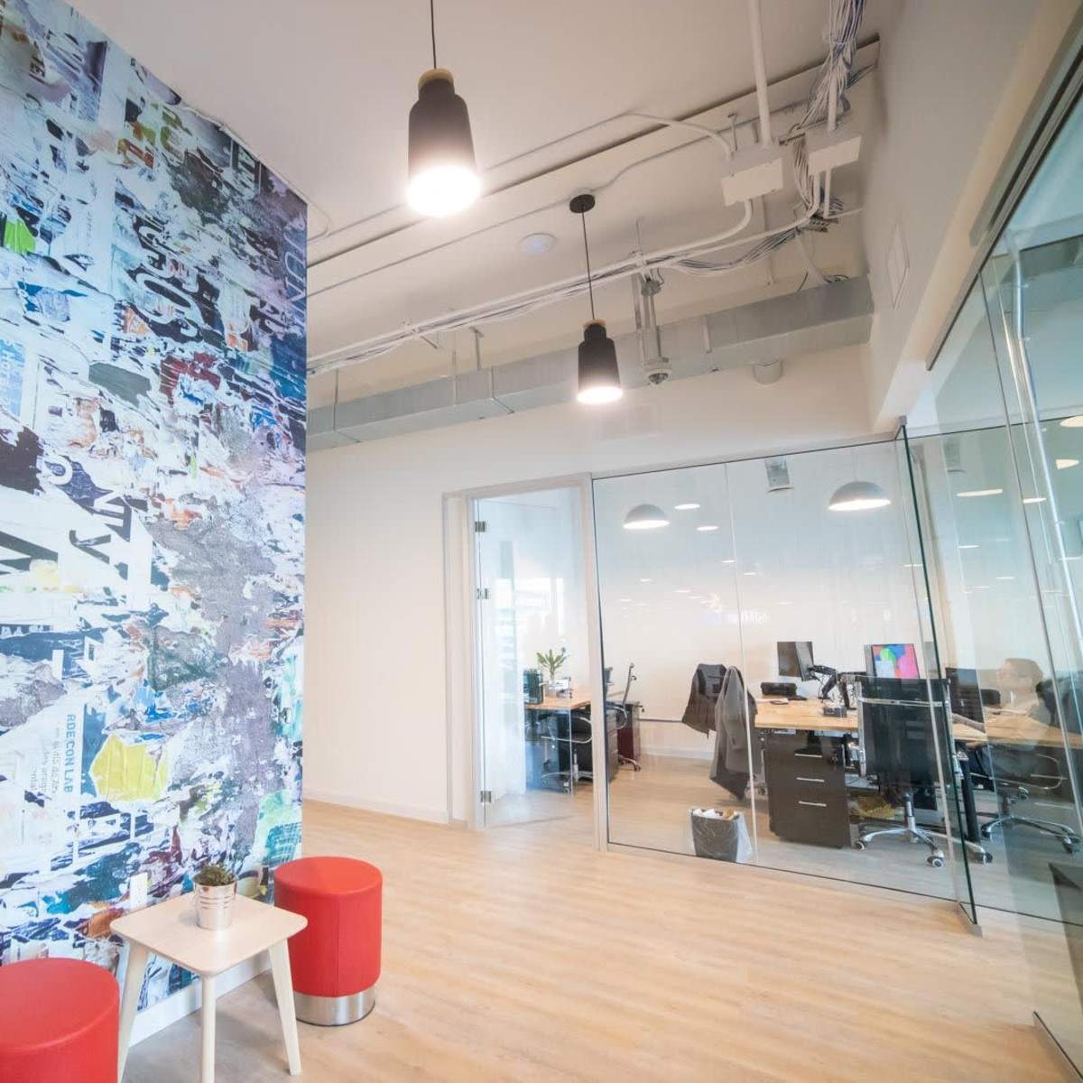 A modern office space featuring a colorful mural on a wall, two red stools, and a glass-walled meeting room in the background.
