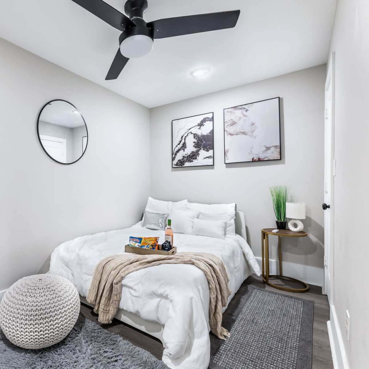 A small bedroom with a bed, mirror, and two framed pictures on the wall, along with a nightstand and a fluffy pouf on the floor.