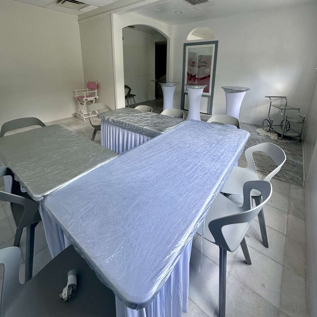 A sparsely furnished room with several tables covered in plastic and surrounded by chairs.