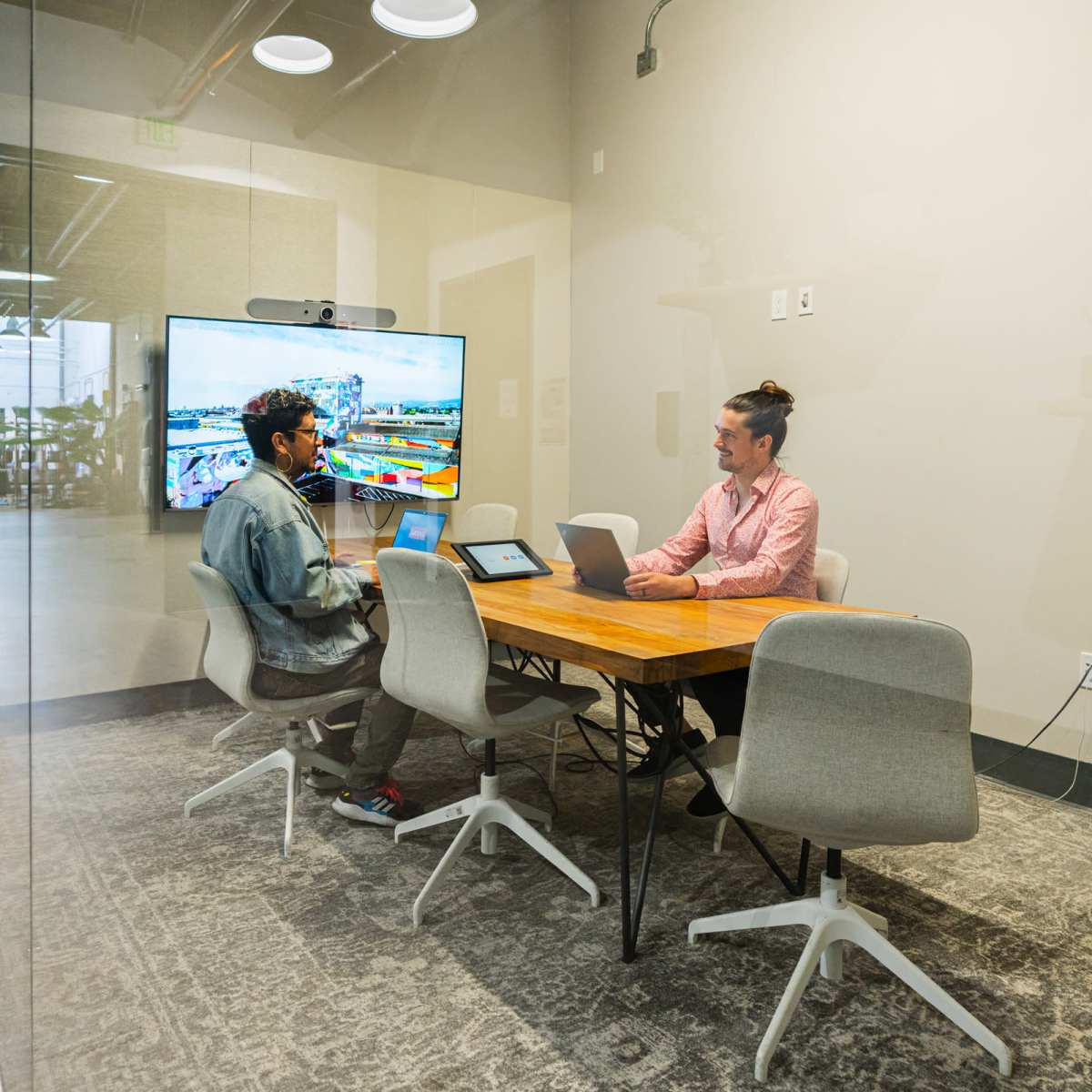 Two people sit at a table in a glass-walled conference room, working on laptops with a large screen displaying a colorful presentation in the background.