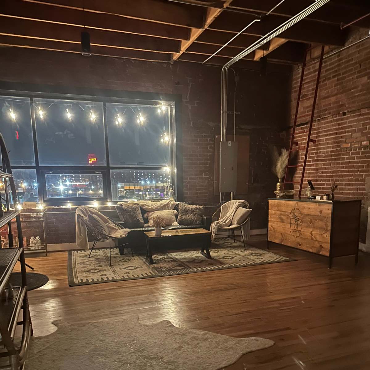 A cozy living space with exposed brick walls, wooden floors, a large window adorned with string lights, and a seating area featuring two chairs and a coffee table.
