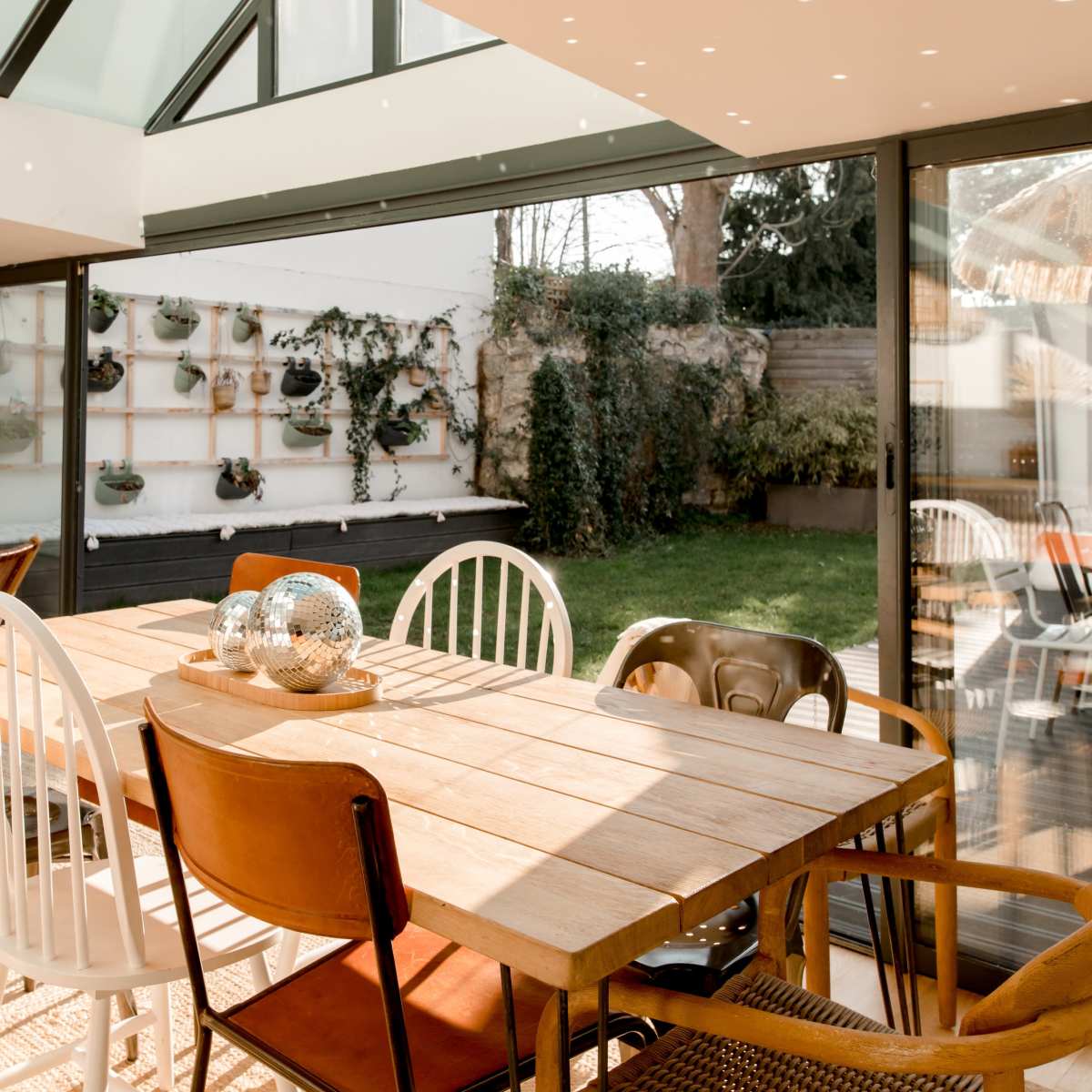 A wooden dining table with various chairs is set in a sunlit room surrounded by glass walls, overlooking a green garden.