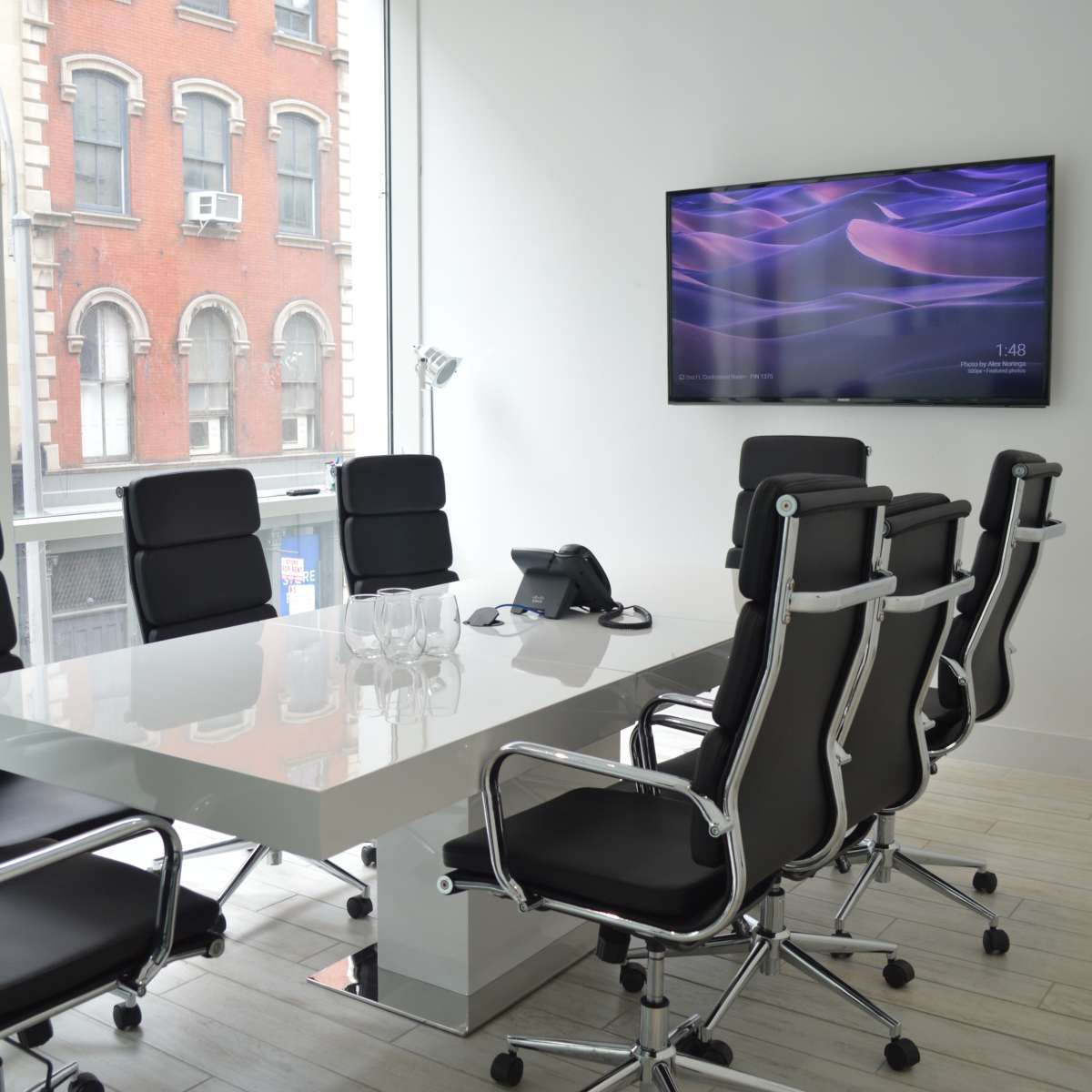 A modern conference room with a large table, eight black chairs, a telephone, and a wall-mounted television, overlooking a city street through a window.