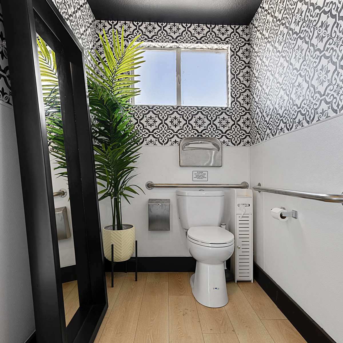 A small bathroom with a toilet, a large mirror, decorative wall patterns, a window, and a potted plant.