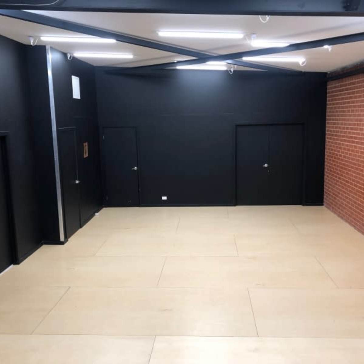 A spacious interior with black walls and two dark doors, alongside a section of exposed red brick wall, illuminated by overhead lights.