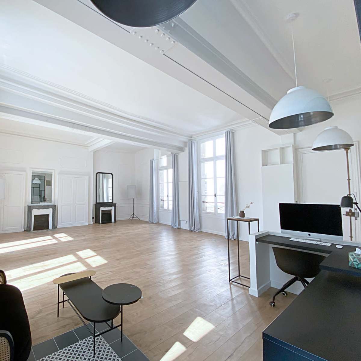 A spacious, sunlit office with light-colored walls, wooden flooring, and large windows, furnished with a black sofa, a glass coffee table, and a desk with a computer.