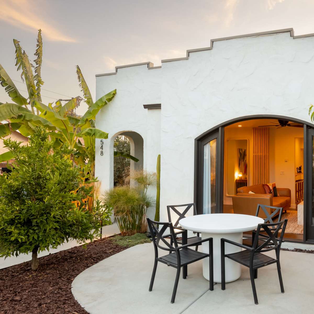 A small outdoor patio features a white table and four black chairs, surrounded by greenery, next to a curved entrance of a white stucco house.