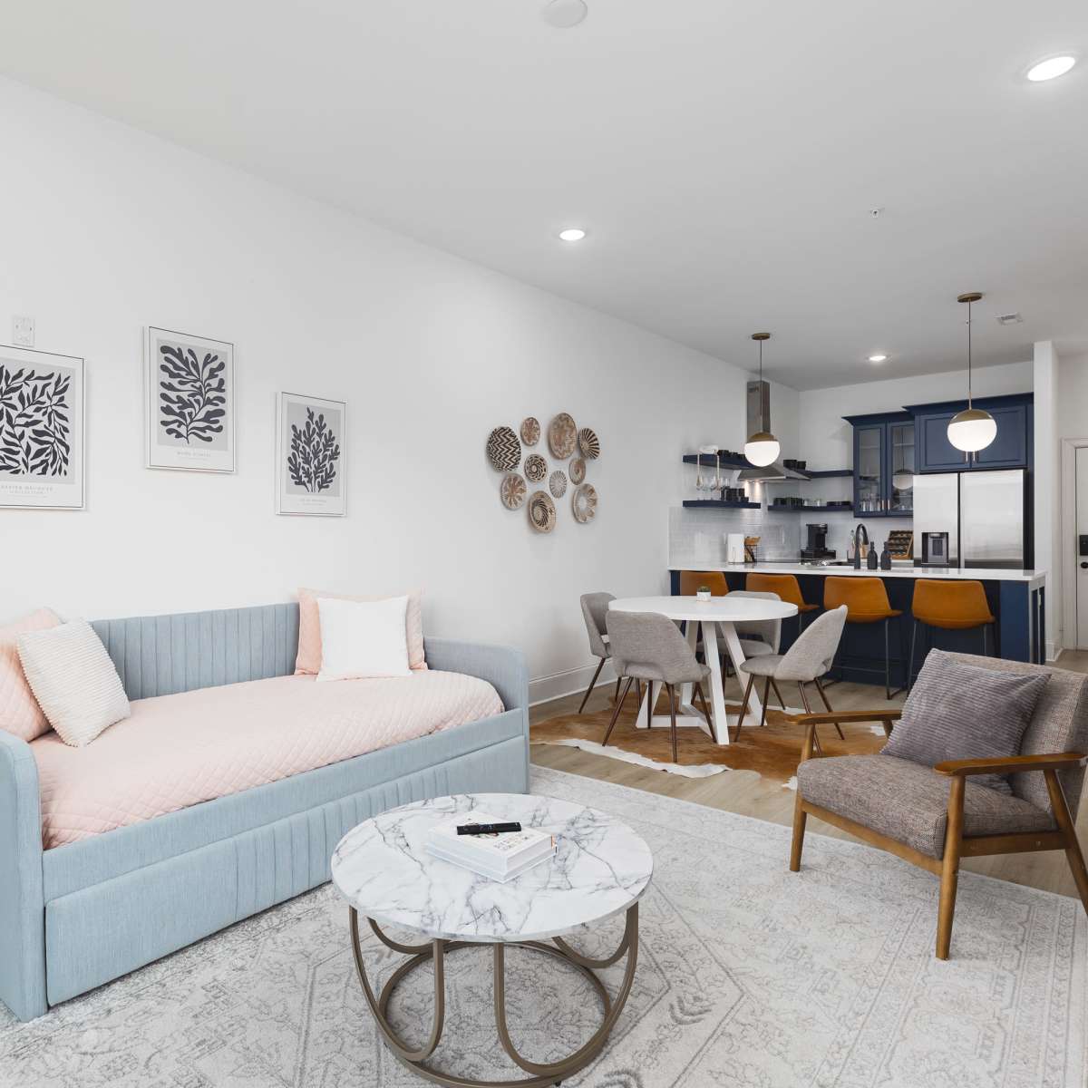 A modern living room with a light blue sofa, a round marble coffee table, and a dining area featuring wooden chairs and a dark blue kitchen in the background.