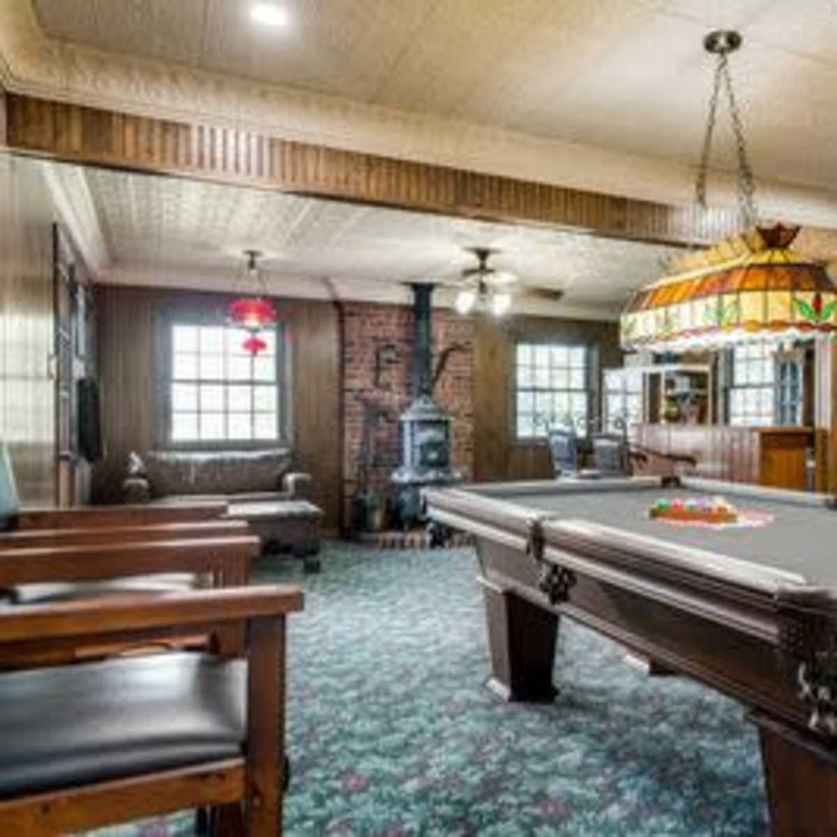 A cozy game room featuring a billiard table, a brick fireplace, wooden paneling, and large windows.