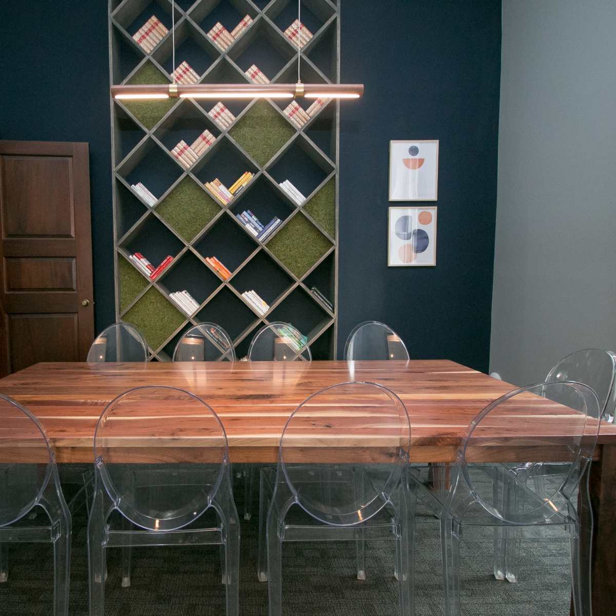 A dining table made of wood is surrounded by clear plastic chairs in a room featuring a bookshelf against a dark wall and two framed artworks.