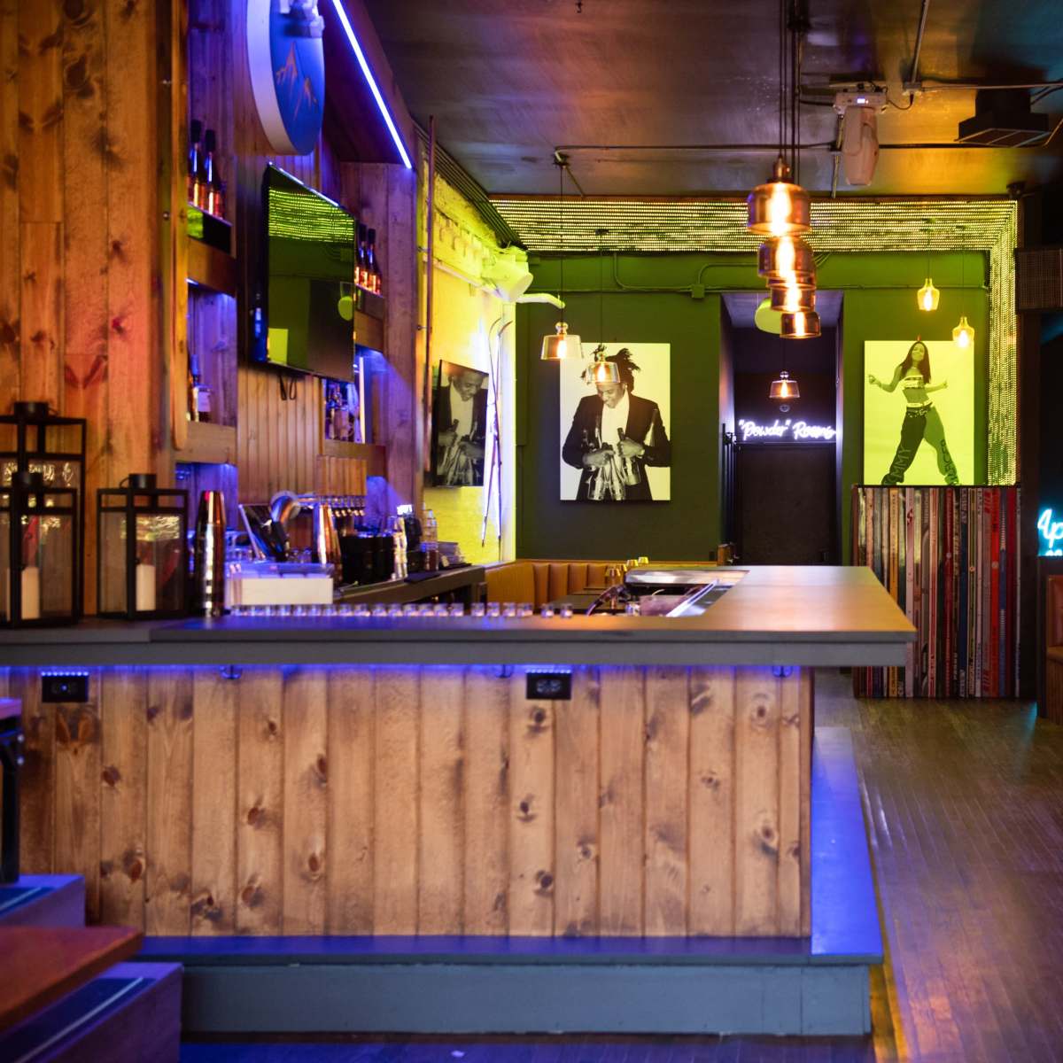 A bar area with a wooden counter, illuminated with colorful lights, and featuring seating along the walls and artwork displayed.
