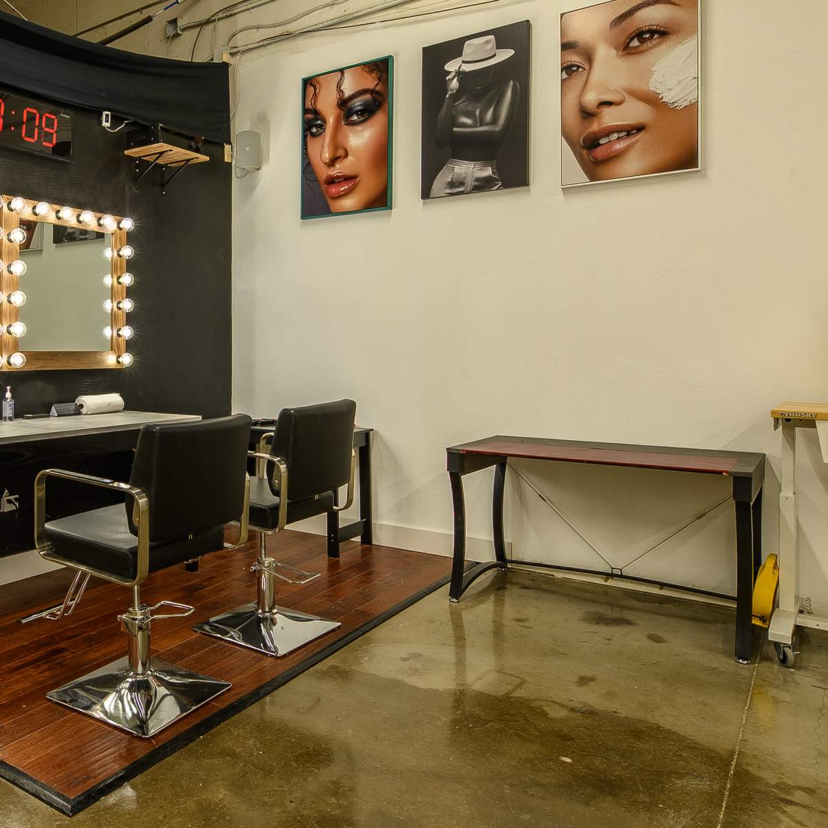 A makeup studio with two chairs, a lighted mirror, and framed photographs on the wall, along with a digital clock displaying the time.