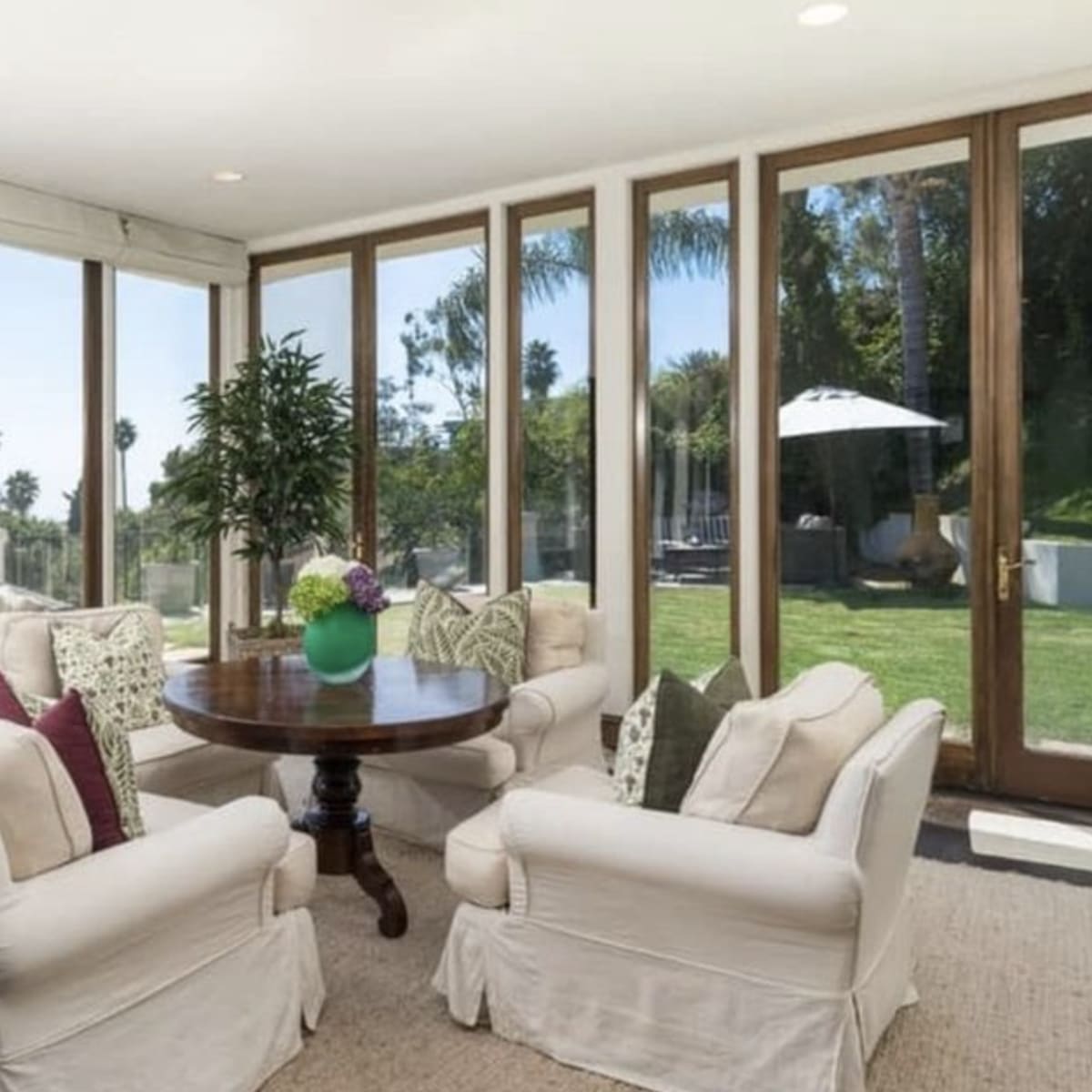 A sunlit living room features large windows, a round table, and beige upholstered chairs arranged around a rug, with views of a garden outside.