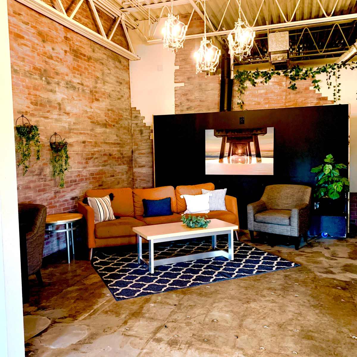 A cozy lounge area featuring an orange sofa, two armchairs, a coffee table, and a decorative rug, all set against a backdrop of exposed brick walls and greenery.