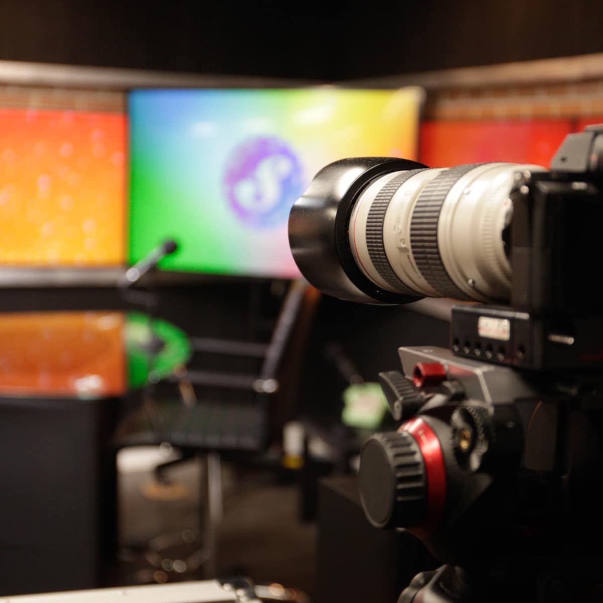 A camera is positioned on a tripod in a broadcasting studio with a colorful backdrop and a monitor displaying a logo.