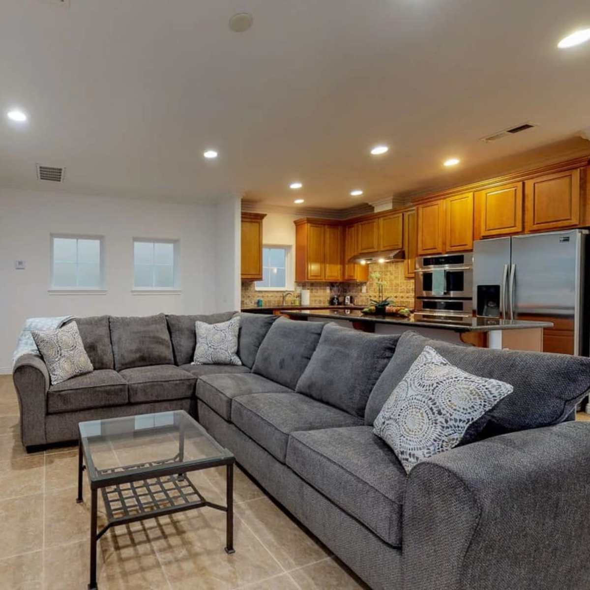 A spacious living room features a gray sectional sofa and a glass coffee table in front of a fireplace, with a kitchen visible in the background.
