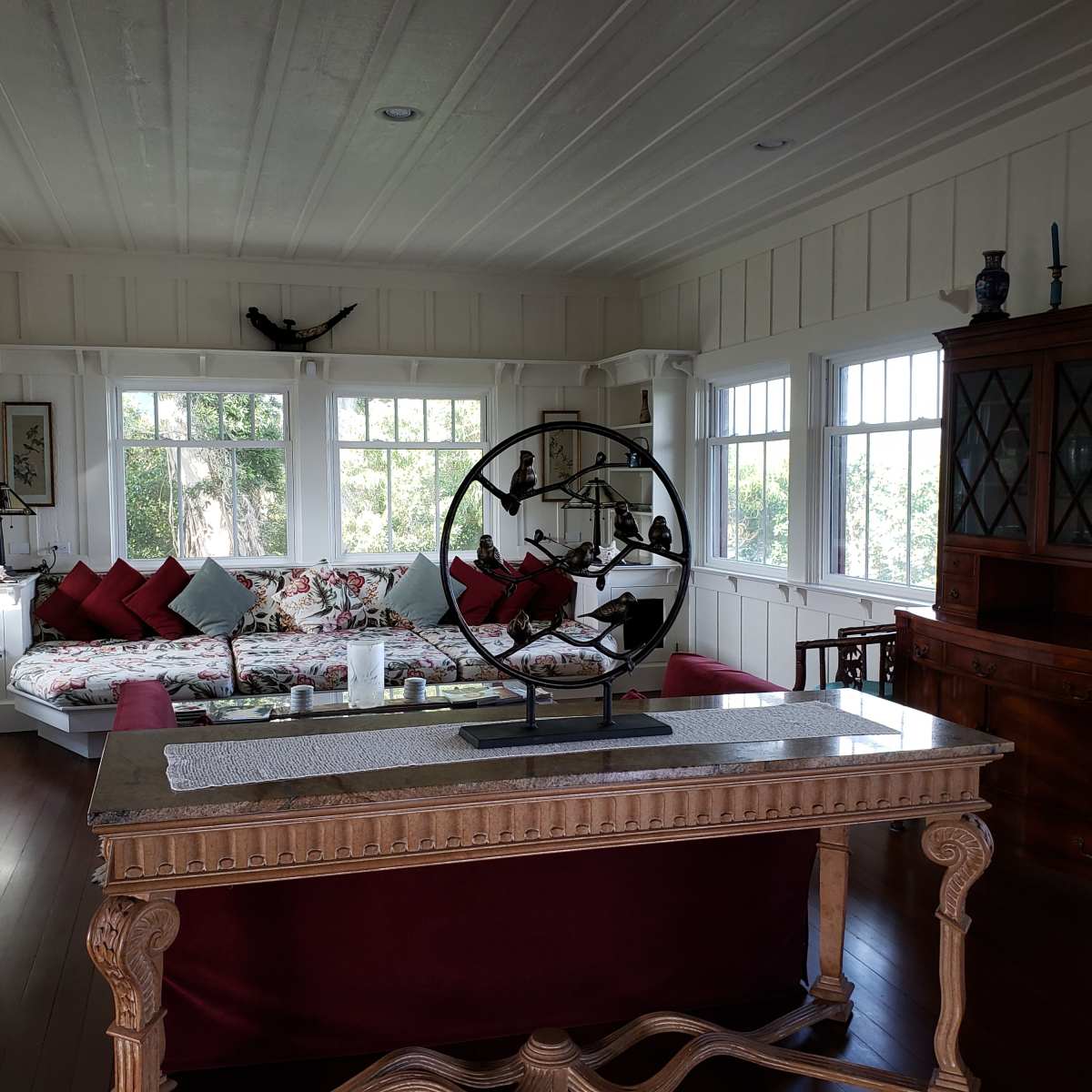 A spacious living room with large windows, a floral-patterned sofa, a wooden dining table, and a decorative display cabinet.