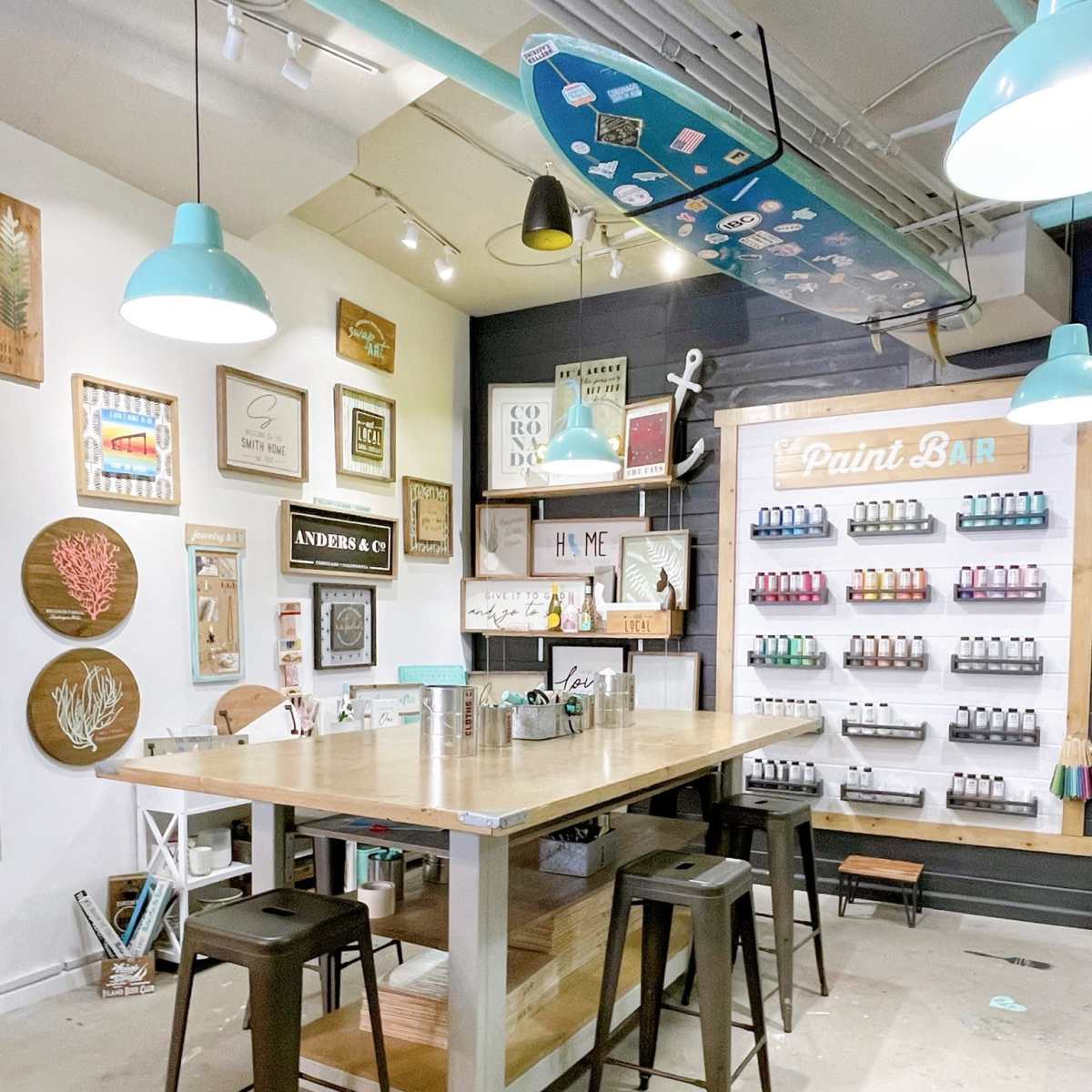 A crafting space featuring a central wooden table surrounded by black stools, with a wall displaying an array of paint bottles and various framed decorations.
