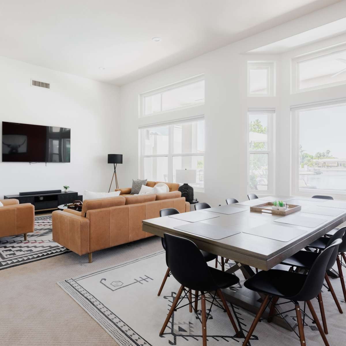 A modern living space features a sectional couch, a dining table with chairs, a large TV mounted on the wall, and tall windows allowing natural light to fill the room.