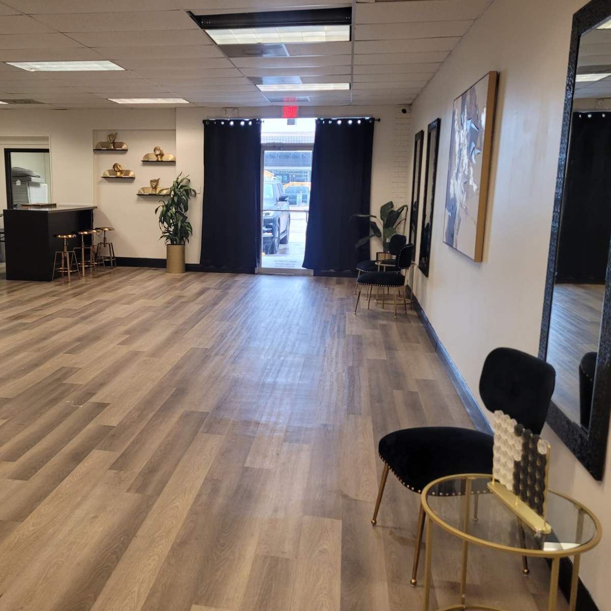 A spacious interior room with wooden flooring, featuring a table and chairs along one wall, a plant, and an open door leading outside.