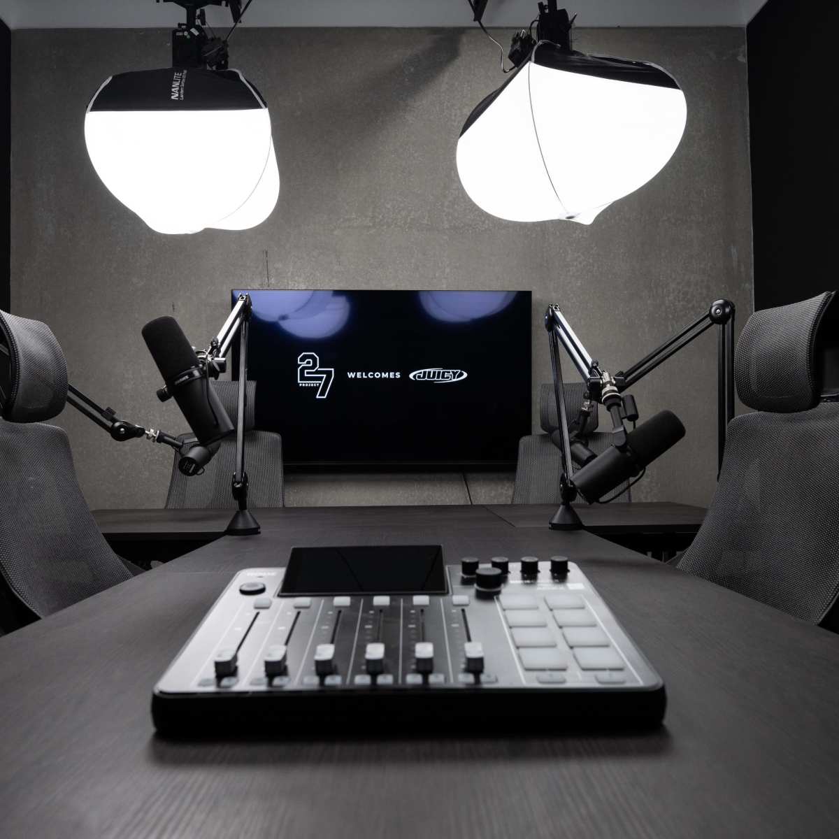 A podcasting studio equipped with two chairs, microphones on adjustable arms, a control panel, and a large screen displaying a welcome message.