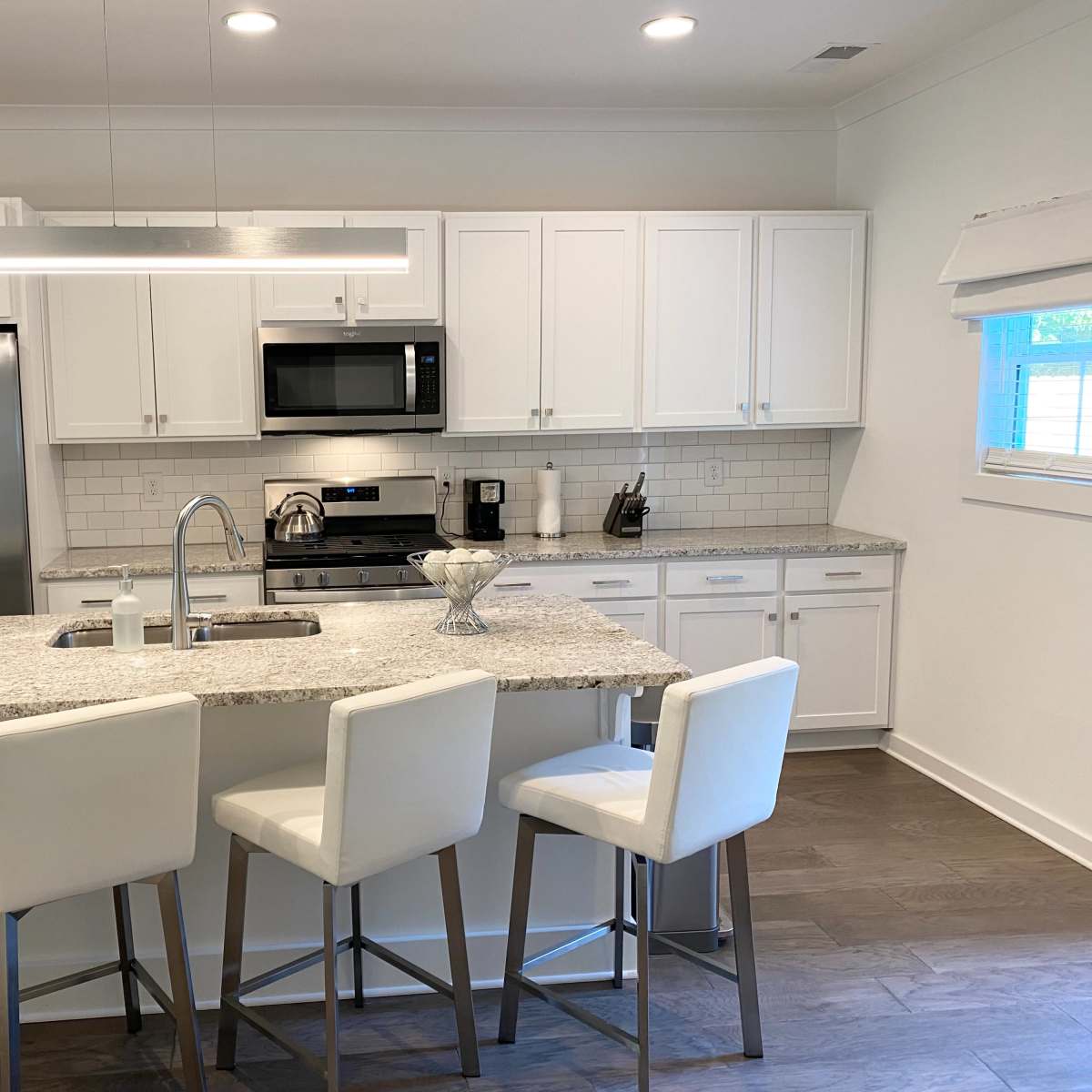 A modern kitchen features white cabinetry, stainless steel appliances, a large granite island with seating, and bright lighting.