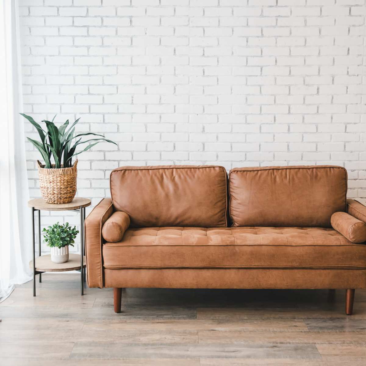 A brown leather couch sits against a white brick wall next to a potted plant on a small table.
