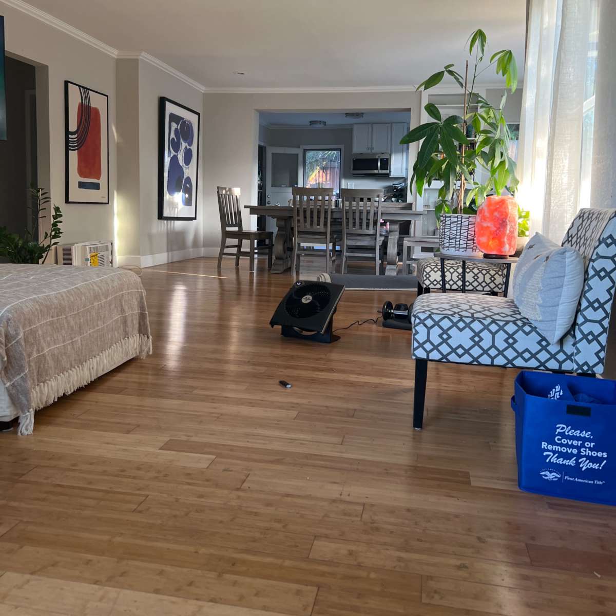 A living room features hardwood flooring, a beige couch, a patterned armchair, a dining table in the background, and a large plant near the window.