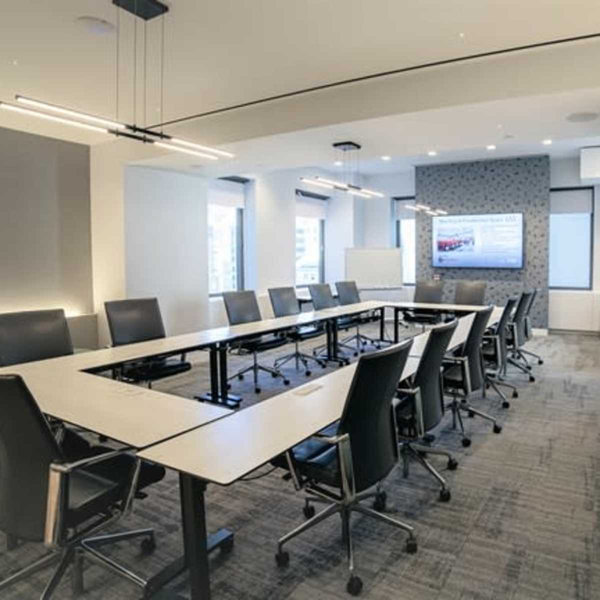 A modern conference room features a long table surrounded by ergonomic chairs, with a large screen mounted on the wall and ample natural light from the windows.