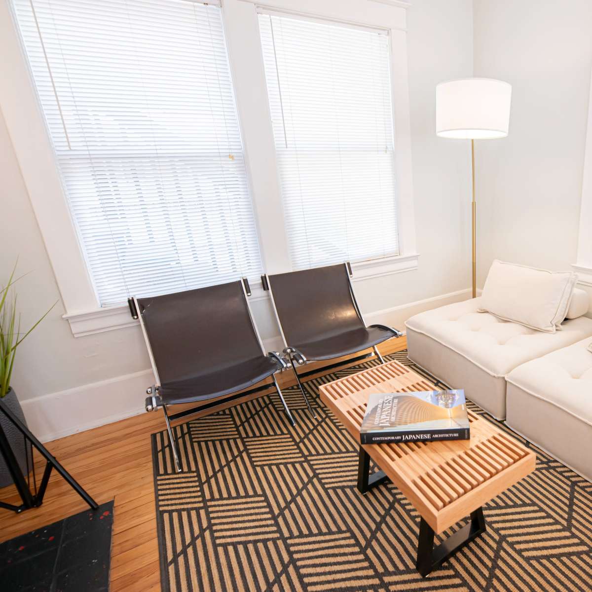 A bright living room with large windows, a sectional sofa, two black chairs, and a wooden coffee table stacked with books.