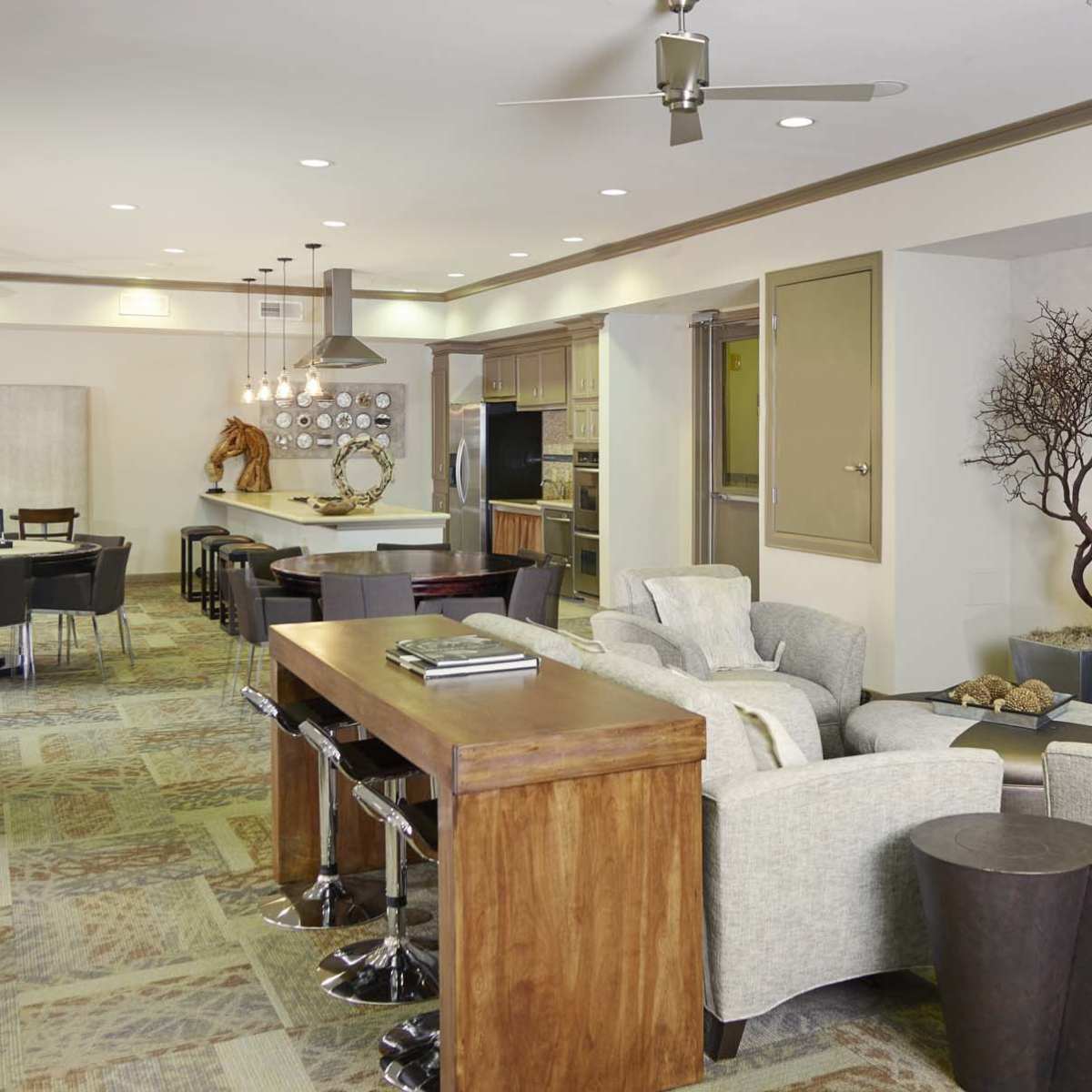 A modern living space with a seating area featuring two light-colored chairs, a wooden bar, and a television mounted on the wall, adjacent to a dining area with a round table and several chairs.